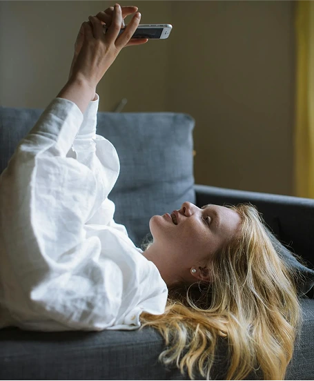 Женщина с распущенными светлыми волосами лежит на диване, подняв телефон для селфи.